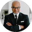 Smiling bald man with glasses wearing a black suit, white shirt, and tie, standing with arms crossed.